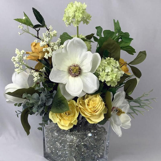 A silk arrangement with artificial flowers of golden roses and ivory magnolias in a square glass vase filled with glass beads, the lifelike artificial foliage blends seamlessly with the flowers. Size: Height 40cm x Width 40cm. Atelier Blooms Auckla