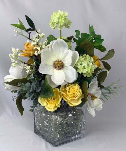 A silk floral arrangement with artificial flowers of golden roses and ivory magnolias in a square glass vase filled with glass beads, the lifelike artificial foliage blends seamlessly with the flowers. Size: Height 40cm x Width 40cm. Atelier Blooms NZ