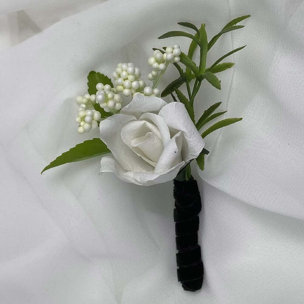 A beautiful handcrafted paper white rose boutonnieres, with a black velvet stem, mixed with artificial foliage, are made to order to match your wedding theme colour and are perfect for the groom, the groomsmen, and fathers for your special occasion. Atelier Blooms